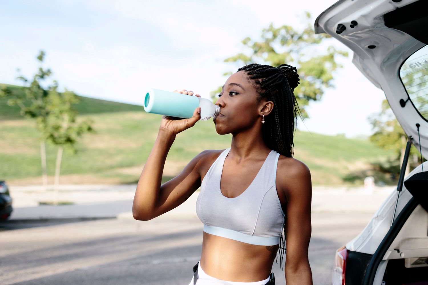 Attractive young black woman on sportswear and with braided hairstyle drinks water next to a car before she starts her training. She's outdoors next to a park in the daytime in a summer morning.