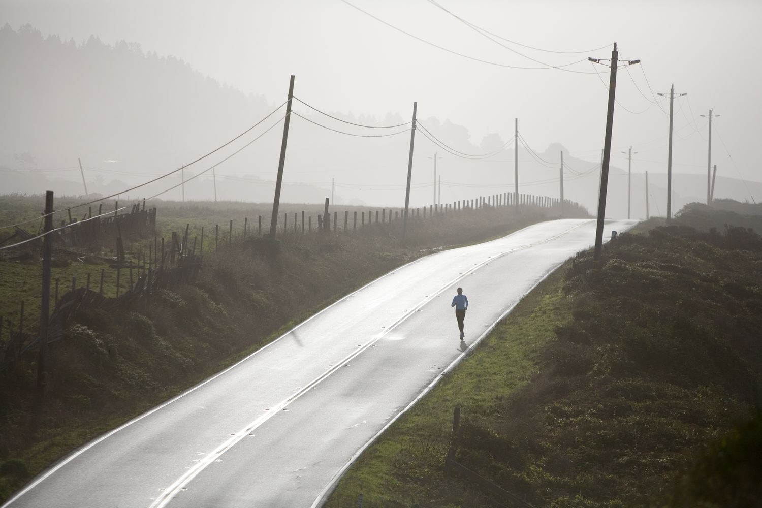 A woman runs in morning fog on deserted road with telephone or electrical poles, northern California coast. (silhouette)