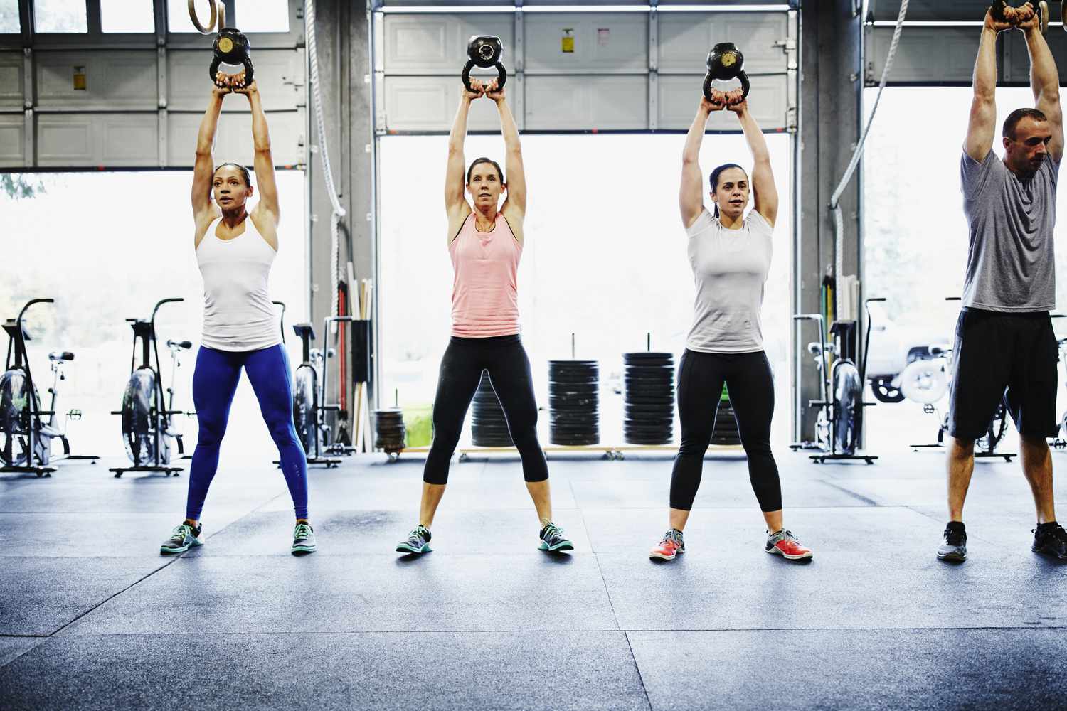 Three women (left) and one man (right) swing kettlebells overhead in a CrossFit gym