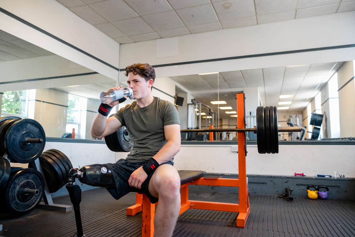 A front view shot of a young teenage male athlete with a prosthetic leg sitting on a gym bench. He is looking forward and drinking water after doing a set of bench presses with a barbell.