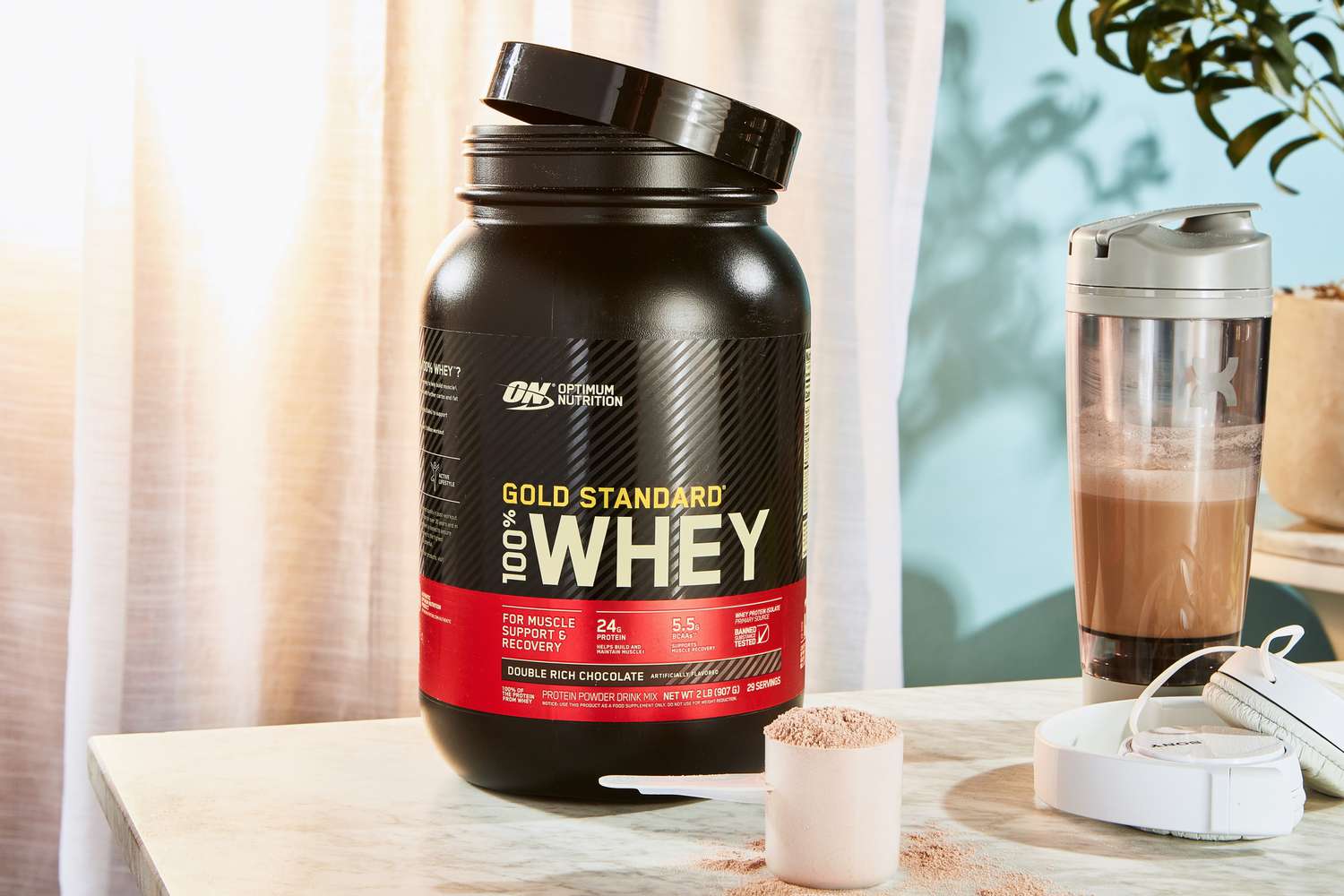 Optimum Nutrition Gold Standard 100% Whey Protein Powder in a scoop next to the jar and blender bottle