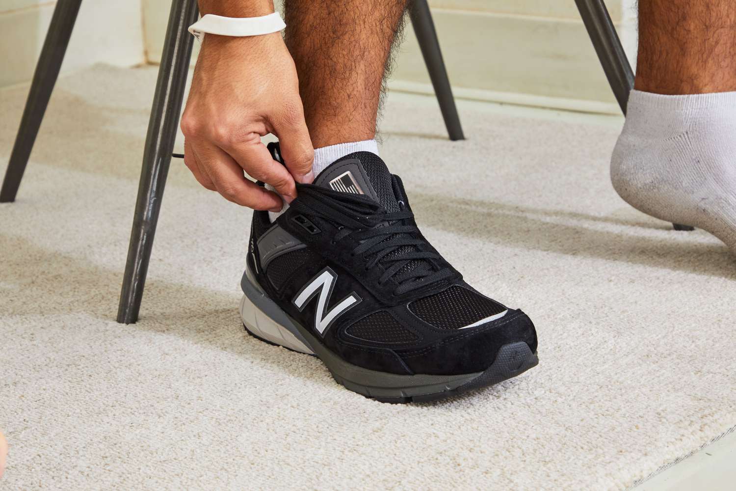 Hand adjusting the laces on the New Balance Men’s M990v5 Running Shoes displayed against a white rug 